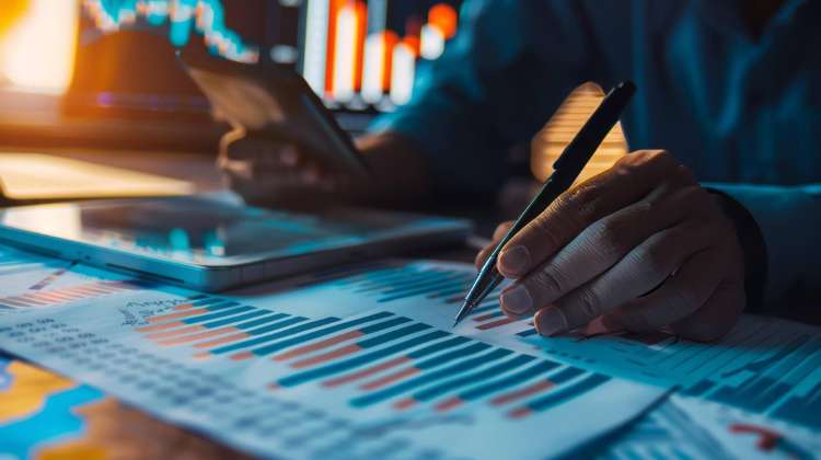 Persona revisando informes financieros y gráficos de datos en una mesa de trabajo, con gráficos en pantalla al fondo.