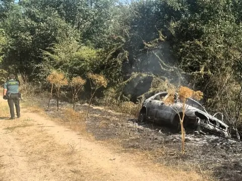 Turismo calcinado tras un accidente mortal en el Camino de los Prodigios, entre Miranda del Castañar y Villanueva del Conde (Salamanca), con un agente de la Guardia Civil inspeccionando la zona
